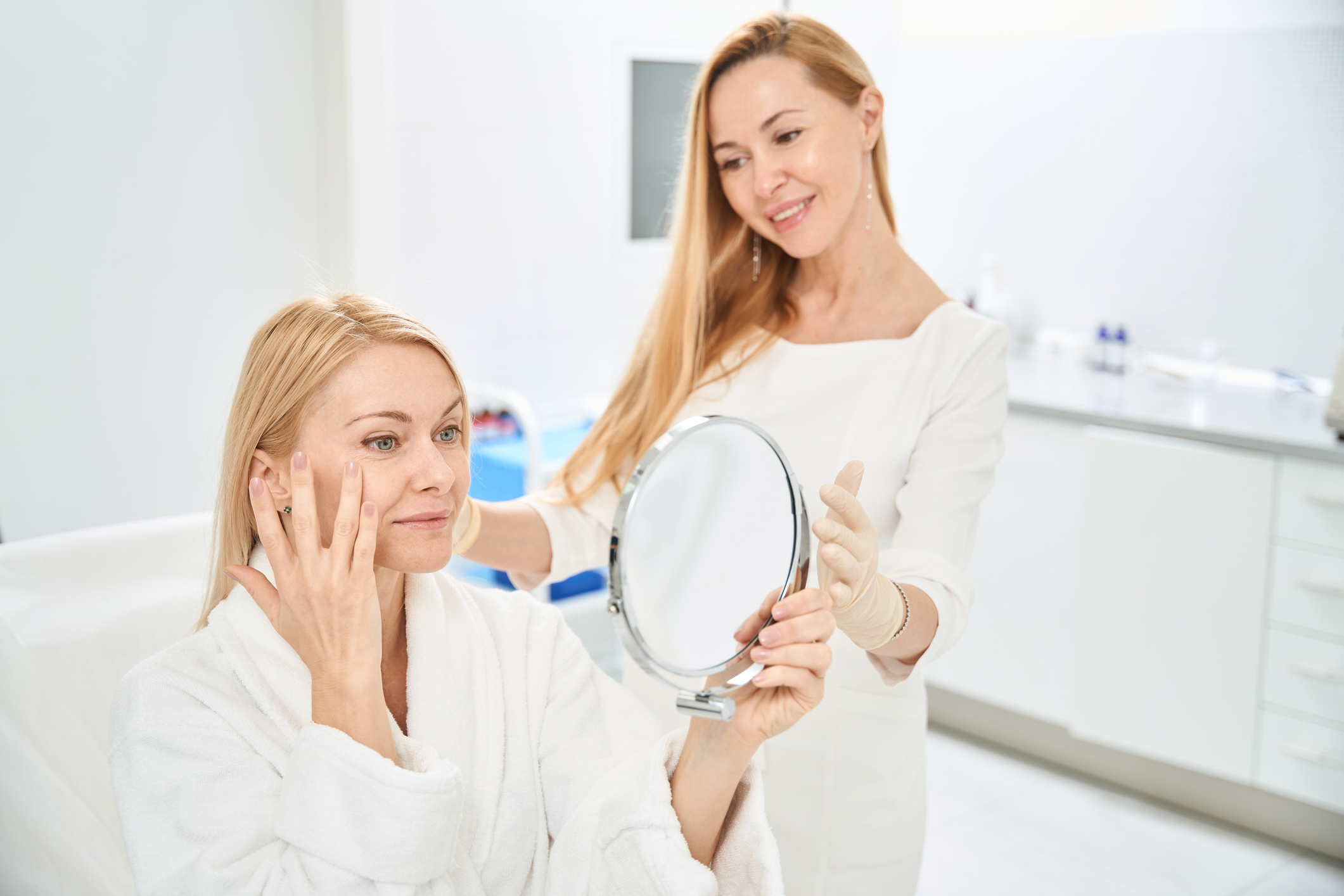 Two women smiling, one holding a mirror while the other touches her face.
