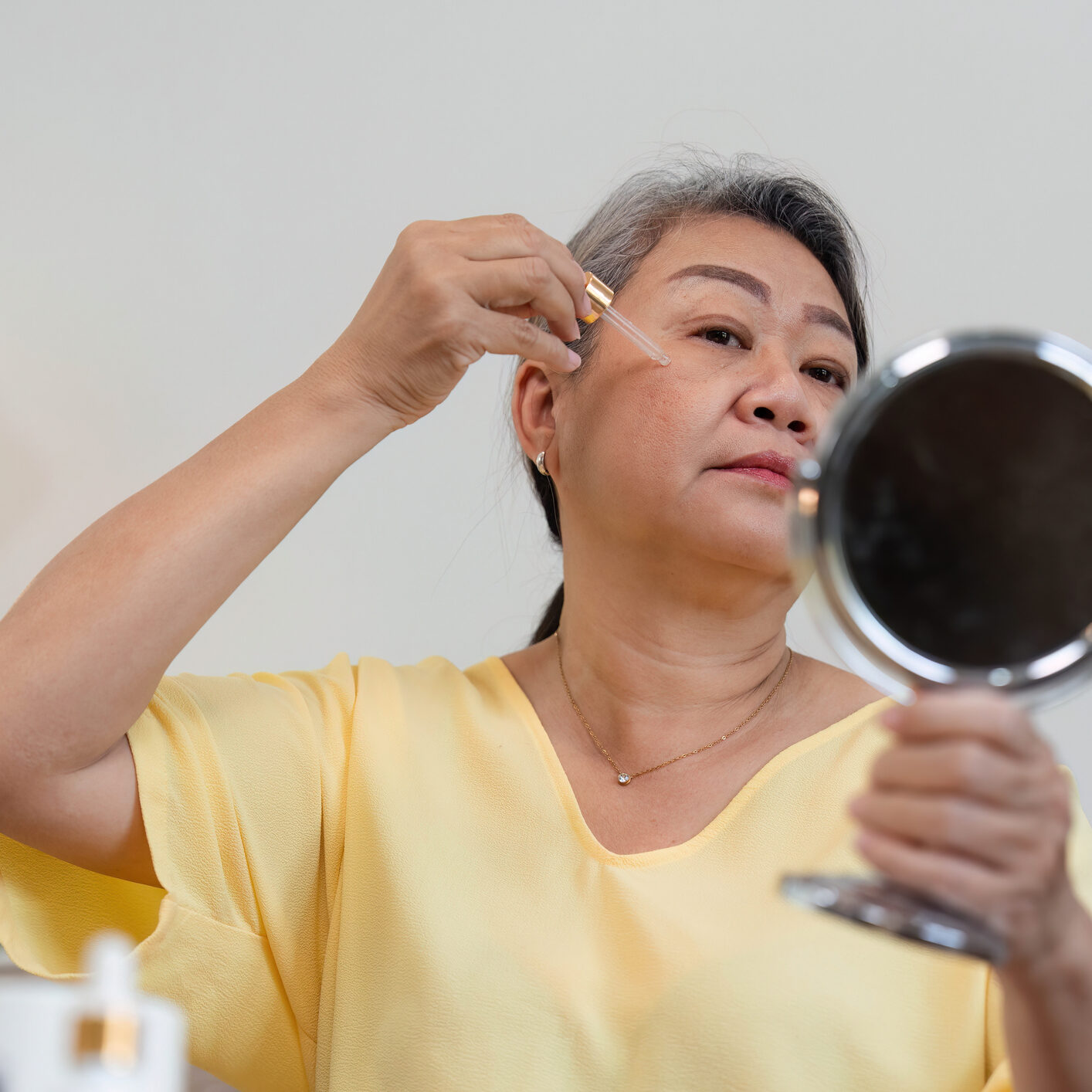 A woman in a yellow shirt applying makeup while looking in a handheld mirror.